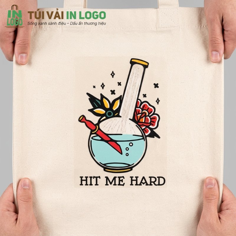 Túi vải tote màu kem được in logo PET nhiều màu sắc nét hình chiếc bong thủy tinh, dao, hoa văn và chữ 'HIT ME HARD', thể hiện chất lượng in ấn trên túi vải.