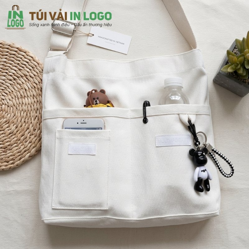Túi vải canvas màu kem đa năng với nhiều ngăn tiện lợi, đựng điện thoại, chai nước, móc khóa gấu, phù hợp làm quà tặng doanh nghiệp bền vững, xu hướng thiết kế 2026.