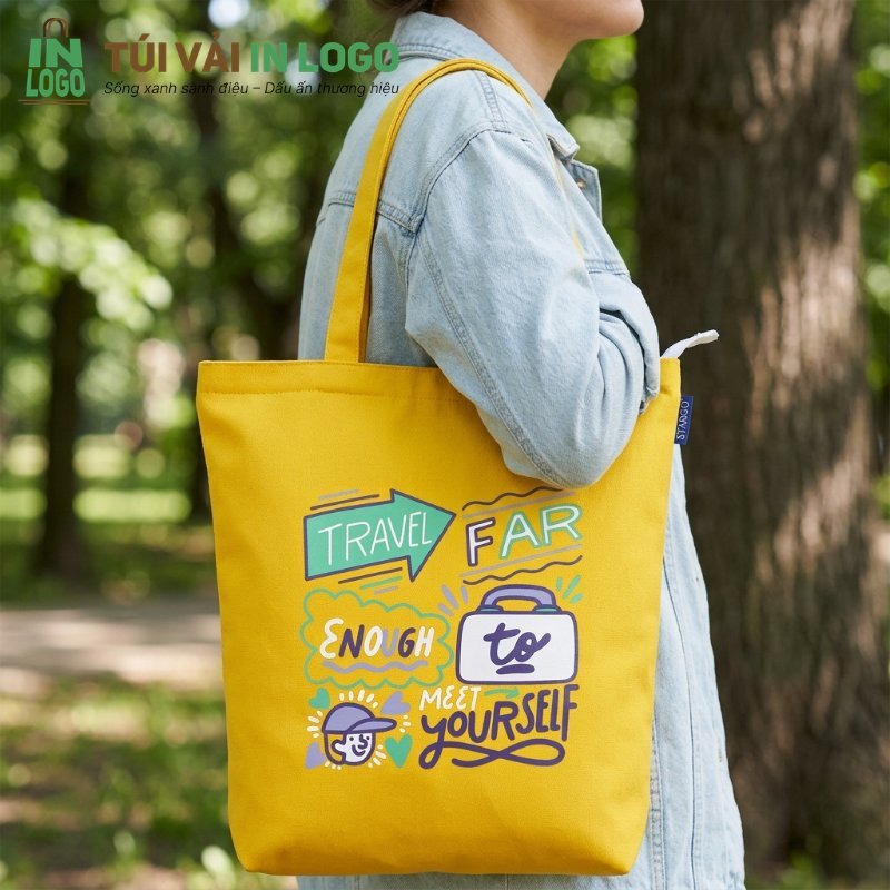 Túi vải canvas màu vàng in logo slogan du lịch "TRAVEL FAR ENOUGH to MEET YOURSELF" đeo trên vai người phụ nữ trong công viên, tượng trưng cho phong cách bền bỉ, thời trang và thân thiện môi trường.